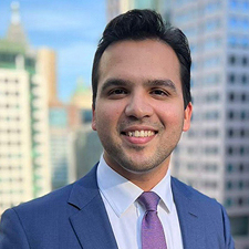 Varun is wearing a suit and smiling. Buildings are visible in the background.