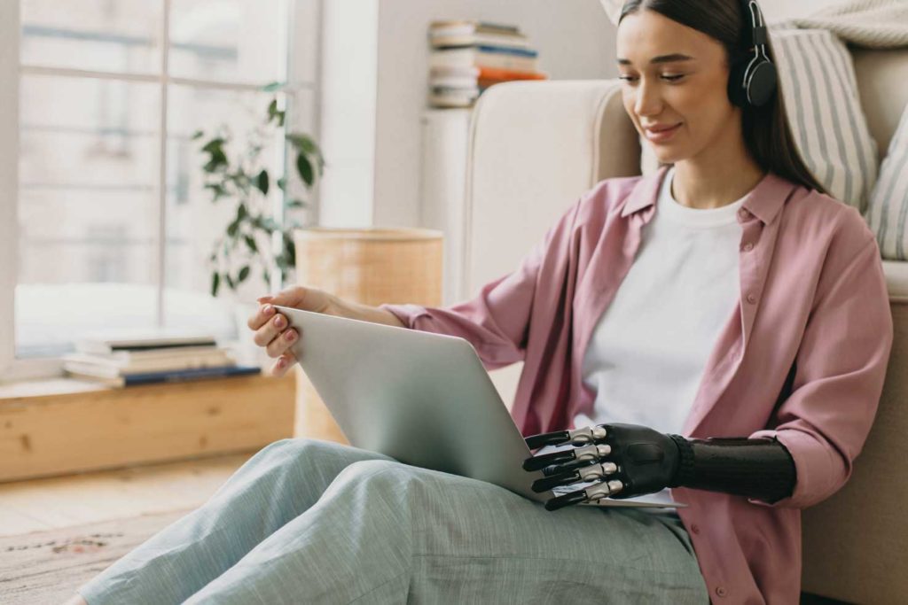A young female with a prosthetic arm sitting on the floor wearing headphones and using a laptop.